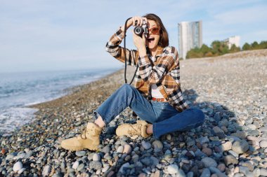 Kayalık bir kıyı şeridinde bir kadın fotoğrafçıyla açık hava portresi çekimi, günlük kıyafetler, sahilde dalgaları ve manzarayı yakalamak.