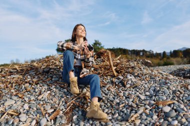 Kayaların ve çakıl taşlarının arasında bir kadın resmi, kot, bot ve ekose gömlekli rahat moda, doğal duruş, açık manzara, zamansız yaşam tarzı anı