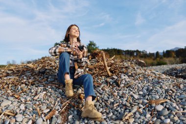 Ekose bluzlu ve botlu bir kadın, elinde kamera, açık gökyüzü altında doğanın tadını çıkarıyor. Kayalık bir sahilde rahat bir yaşam tarzı portresi. Sahne sükuneti, macerayı ve dışarıyı vurguluyor.