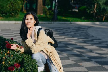 Açık hava parkında canlı çiçeklerin yanında gülümseyen kadın, rahat bir stil, doğa ve gündüz keyfi sergiliyor, açık havada huzurlu bir anı yakalıyor..