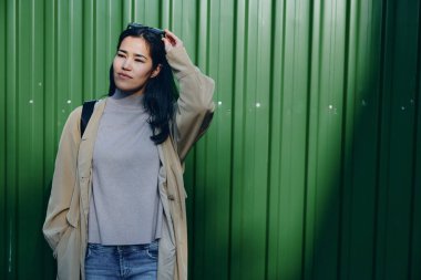 Örgü süveter ve açık renk ceket giyen sıradan bir kadın yeşil bir duvarın yanında poz veriyor, şehir modası havası ve sakin bir ifade, modern sokak tarzı ve çok yönlü gardırobu yakalıyor.