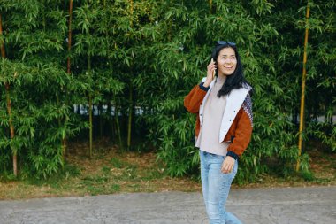 Dışarıdan gülümseyen kadın telefonla arıyor, şık dış giyim ve kotlar, yeşil ve bambuların arasında yakalanmış, doğal manzara ve şehir havasında sıradan bir yaşam tarzı.