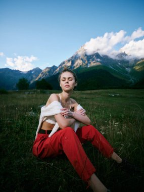 Açık hava sahnesi, tarlada rahat bir kadın figürü, kalın kırmızı pantolon ve rahat bluz, sakin doğa hali, dağlık manzara ve yukarıdaki uzak tepeler.