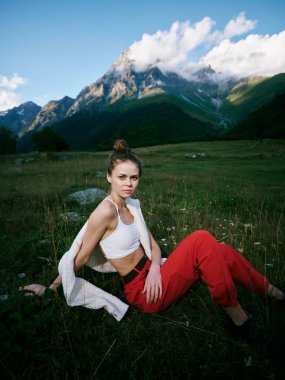 Açık hava sahnesi, yeşil çimlerde dinlenen genç bir kadın, yukarıdaki dağ kulesi, günlük kıyafetler, beyaz atlet bluz ve kırmızı pantolon, sakin manzara ve parlak gün ışığı..