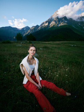 Çimenli bir çayırda oturan bir kadını gösteren manzara, koyu kırmızı pantolon ve beyaz üst, dramatik alp dağları ve geniş gökyüzü, modadan etkilenen sakin manzara