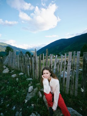 Alp arazisinde çitlere yaslanan bir kadının olduğu açık hava sahnesi, manzaralı dağlar, çimenler ve kayalar, gizli moda çekimi, doğa havası ve sakin kırsal kesimde seyahat tarzı.
