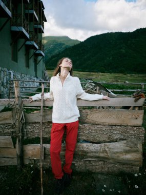 Açık hava sahnesinde tahta bir bariyerin yanında bir kadın, arka planda dağlar, beyaz gömlek ve kırmızı pantolon, sükunet, keşif ve doğa ile bağlantı..