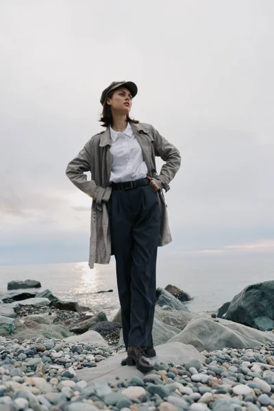 Gündelik kıyafetli genç bir kadın, elleri kalçasında çakıl taşı plajında poz veriyor. Bulutlu gökyüzü ve sakin su arka planı doğal ortamdaki gevşemiş ruh halini ve modern tarzı vurguluyor.