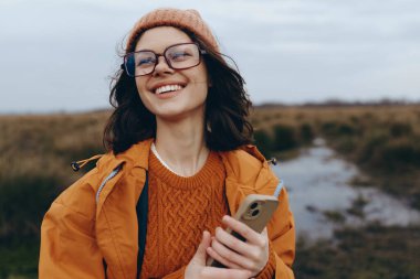 Gözlüklü ve turuncu giysili mutlu bir kadın açık havada dururken akıllı telefon tutuyor. Sonbahar modasını, teknoloji kullanımını ve neşeli ruh halini vurgulayan yaşam tarzı fotoğrafı.