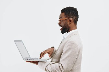 Kısa kıvırcık saçlı ve sakallı, açık renk ceket giyen profesyonel çalışma ortamında dizüstü bilgisayar kullanan kendine güvenen genç bir adam. Parlak beyaz arkaplan, teknoloji, iş ve modern alanlara odaklanıyor