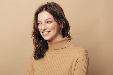 Tarafsız arkaplanda duran bej balıkçı yaka kazaklı, kıvırcık saçlı mutlu kadın portresi. Doğal güzellik kavramı, gündelik moda ve rahatlamış pozitif ifade..