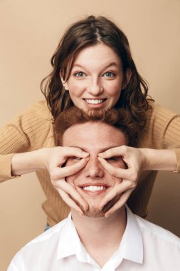 Erkeğin arkasındaki gülümseyen kadın, kalbi yüzünde elleriyle şekillendirir. Gayri resmi giyinen genç çift bej arka planda poz veriyor. Şefkat, neşe ve şakacı etkileşim kavramı.