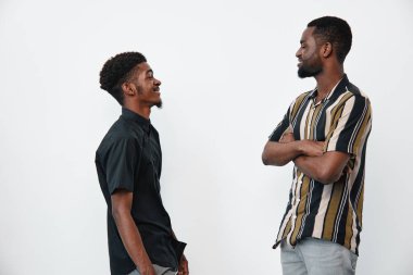 Siyah tenli, sıradan gömlekli Afro-Amerikalı erkekler yüz yüze durup gülümsüyor. Beyaz arka planda dostluk, iletişim ve olumlu etkileşim.