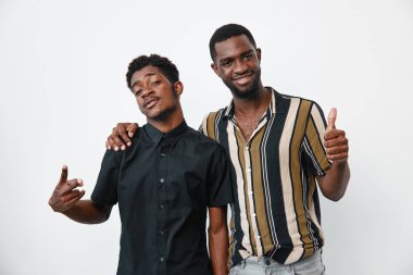 Siyah tenli Afro-Amerikalı erkekler birbirlerine yakın durup gülümsüyor ve beyaz arka planda el işaretleri yapıyor. Arkadaşlık, gündelik tarz, pozitif ruh hali ve bağlantı.