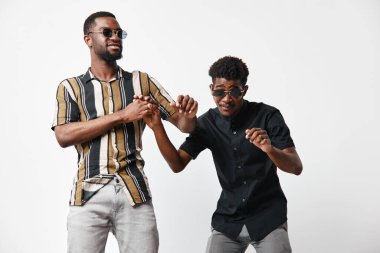 Gündelik kıyafetleri ve güneş gözlükleriyle dans eden ve beyaz arka planda kahkahalar atan iki siyah adam. Genç arkadaşlar içeride eğleniyor ve mutluluklarını ifade ediyorlar..