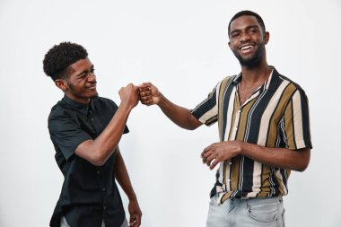 Koyu tenli, sıradan kıyafetleri olan, gülümseyen ve beyaz arka planda yumruk tokuşturan genç siyahi adamlar. Erkek arkadaşlar arasında dostluk, bağlantı ve neşeli anlar..
