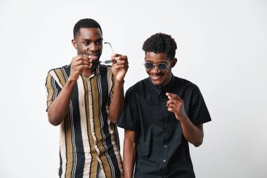Siyah tenli, güneş gözlüklü ve beyaz arka planda günlük giysiler giyen iki siyah adam. Arkadaşlık, moda, mutluluk ve yaşam tarzı. Stüdyoda neşe ve havalı bir tarz..