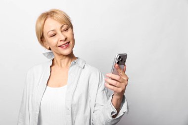 Kısa sarı saçlı, akıllı telefon tutan ve beyaz arka planda gülümseyen mutlu orta yaşlı kadın. Dijital iletişim, teknoloji, yaşam tarzı ve modern sosyal etkileşim kavramı.