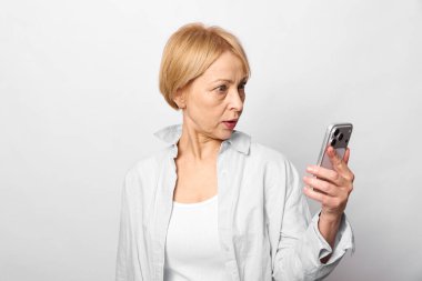 Kısa saçlı, beyaz gömlekli olgun bir kadın akıllı telefon tutuyor ve şaşırmış görünüyor. Beyaz arka planda izole edilmiş. Teknoloji kavramı, iletişim, beklenmedik reaksiyon, dijital cihaz.