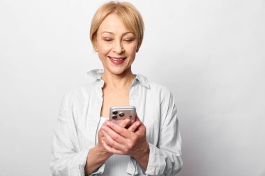 Beyaz tişörtlü, kısa sarı saçlı mutlu orta yaşlı bir kadın, arka plandaki ışığa bakarken iki eliyle akıllı telefonu tutuyor ve gülümsüyor. Teknoloji ve iletişim konsepti.