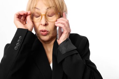 Gözlüklü ve siyah ceketli orta yaşlı bir kadın telefonu kulağına dayayıp endişeli bir ifadeyle gözlerini kapatıyor. Beyaz arka plan, iletişim ve duygu konsepti üzerine stüdyo çekimi.