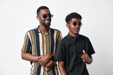 Siyah tenli, sıradan giyinmiş ve güneş gözlüklü Afro-Amerikalı erkekler. Parlak stüdyo ortamında gülümseyen ve poz veren iki genç arkadaş..