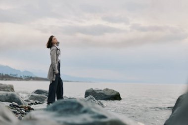 Palto giyen kadın kayalık sahilde bulutlu gökyüzü ile sakin okyanusa bakıyor. Huzurlu sahne yalnızlığı, yansımayı ve doğayla olan bağlantıyı su aracılığı ile aktarır..