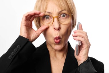 Şok olmuş gözlüklü ve kısa sarı saçlı bir kadın cep telefonunu kulağına yakın tutuyor, diğer eliyle gözlüğü ayarlıyor. Sürpriz, iletişim, teknoloji ve duygusal tepki kavramı.