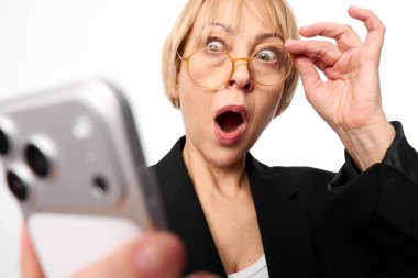 Gözlüklü ve siyah ceketli sarışın kadın elinde akıllı telefon tutarken şaşkınlıkla tepki veriyor. Geniş gözleri ve ağzı, beyaz arka plana karşı şaşkınlığını ifade ediyor..