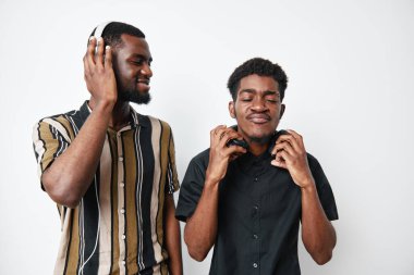 Koyu tenli Afrikalı erkekler kulaklıkla müzik dinliyor, gülümsüyor ve beyaz arka planda günlük gömleklerle dikiliyorlar. Rahatlama, dostluk ve yaşam tarzı anı.