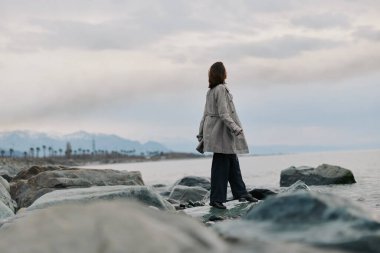 Ekose ceket giyen kadın bulutlu gökyüzünün altında deniz kenarındaki kayalarda duruyor, suya bakıyor. Görüntü, okyanus kıyısındaki huzur dolu yalnızlığı, açık havayı ve yansıtıcı havayı yakalar..