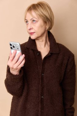 Kahverengi desenli ceketli orta yaşlı sarışın kadın akıllı telefon tutuyor ve bej arka planda yan bakıyor. İletişim, teknoloji, günlük yaşam tarzı ve modern yetişkin portresi kavramı.
