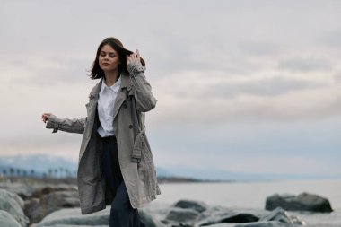 Sıradan palto giyen genç bir kadın bulutlu arka planda sakin deniz kenarında kayalık sahilde yürüyor. Doğa, moda, özgürlük, açık hava yaşam tarzı ve huzurlu ruh hali kavramı..