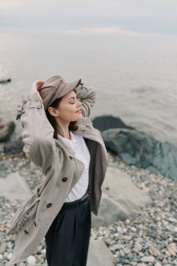 Açık renk ceketli ve şapkalı sakin genç kadın kayalık sahilde duruyor gözleri kapalı, temiz deniz havasını hissederek. Doğa kavramı, rahatlama, özgürlük ve okyanus yakınlarındaki huzurlu kıyı yaşamı..