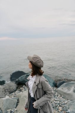 Kayalık sahilde dokulu palto ve şapka giyen ve sakin okyanusa bakan kadının portresi. Bulutlu hava sakin atmosferi güçlendirir. Düşünce, moda ve doğal manzara teması.