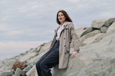 Bej renkli trençkot ve koyu renk pantolon giyen genç bir kadın bulutlu bir arka plan ile kıyı kayalıklarında oturuyor. Rahat duruş ve doğal görünüm sakinliği ve açık hava tarzını yansıtıyor..