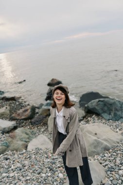 Palto ve bere giyen gülümseyen kadın arka planda sakin denizle kayalık sahilde poz veriyor. Neşeli açık hava anları, doğal rahatlama, gündelik moda ve deniz kenarı mutluluğu kavramı.