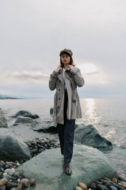 Yün ceket ve şapka giyen şık bir kadın kayalık sahilde okyanus manzaralı pozlar veriyor. Bulutlu gökyüzü ve doğal manzara gelişigüzel sonbahar modasını ve sakin atmosferi vurguluyor.