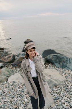 Bej ceket ve desenli şapka giyen gülümseyen kız arka planda sakin bir okyanusla çakıl taşı plajında duruyor. Gündelik sonbahar kıyafetleri, doğa ve deniz kenarında rahat bir hava..
