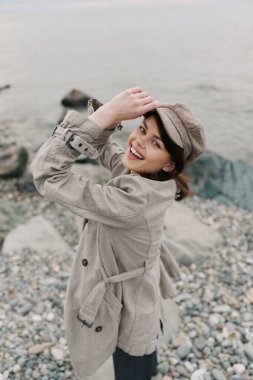 Kumaşlı palto ve kep giyen mutlu kadın arka planda deniz olan çakıl taşı sahilinde duruyor. Günlük kıyafet ve doğal ortam rahat ve neşeli bir ortam yaratır..