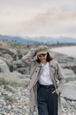 Gülümseyen kadın gri desenli ceket ve uyumlu şapka takıyor uzak dağlarla ve bulutlu gökyüzüyle kayalık kıyı boyunca geziniyor. Rahat bir hava, sonbahar modası ve kıyı manzarası açık havada çekildi..