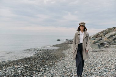 Bej ceketli ve şapkalı kadın sakin denizin kıyısındaki çakıl taşı plajında sessizce yürüyor. Bulutlu gökyüzü ve kayalık kıyı doğadaki sakin, yansıtıcı atmosfere eklenir.