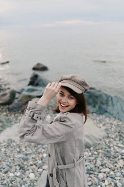 Moda ceketli ve şapkalı mutlu kadın okyanus arka planlı çakıl taşı sahilinde duruyor. Yumuşak doğal ışıklandırma neşeli havayı ve sonbahar kıyafet tarzını arttırır. Deniz kenarında rahatlama ve moda kavramı.