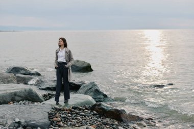 Kayalık kıyı şeridinde sıradan kıyafetli genç bir kadın sakin okyanusa bakıyor. Yumuşak ışık ve bulutlarla kasvetli bir atmosfer. Yalnızlık, yansıma ve doğal manzara kavramı..