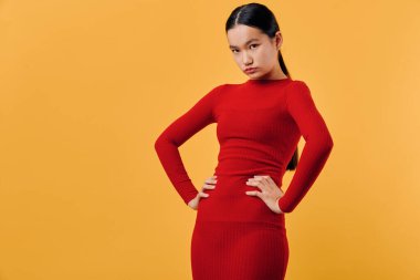 Stüdyoda elleri kalçasında, parlak turuncu arka planı olan kırmızı elbiseli Asyalı bir kadın. Moda portresi, kendine güveni olan poz, günlük kıyafetler ve saç stili.
