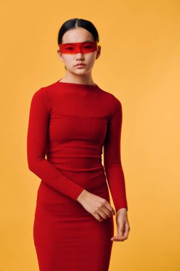 Dar kırmızı elbise giyen şık bir kadın ve sarı arka planda kırmızı şeffaf gözlükler takıyor. Moda fotoğrafçılığı, çağdaş tarz, cesur renkler, zarif ve kendinden emin ifade.