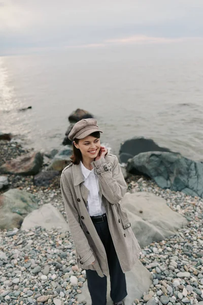 Bej ceket ve desenli şapka giyen gülümseyen kız arka planda sakin bir okyanusla çakıl taşı plajında duruyor. Gündelik sonbahar kıyafetleri, doğa ve deniz kenarında rahat bir hava..