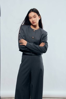 Koyu gri takım elbiseli kendine güvenen Asyalı kadın modeller moda ve profesyonellik için moda fotoğraf çekimlerinde kollarını kavuşturup hafif arkaplanlara karşı poz veriyorlar..