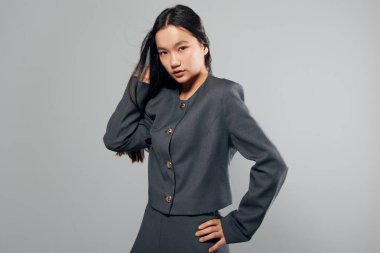 Uzun siyah saçlı, koyu renk ceketli ve etekli, gri arka planda, elleri belinde, moda tarzında duran kendine güvenen genç bir kadının portresi..