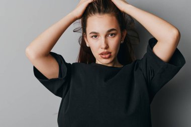 Siyah saçlı, siyah bluzlu, elleri saçında genç bir kadının portresi. Gri arkaplan önünde duran kadın model odaklanmış görünüş ve gündelik tarzda.